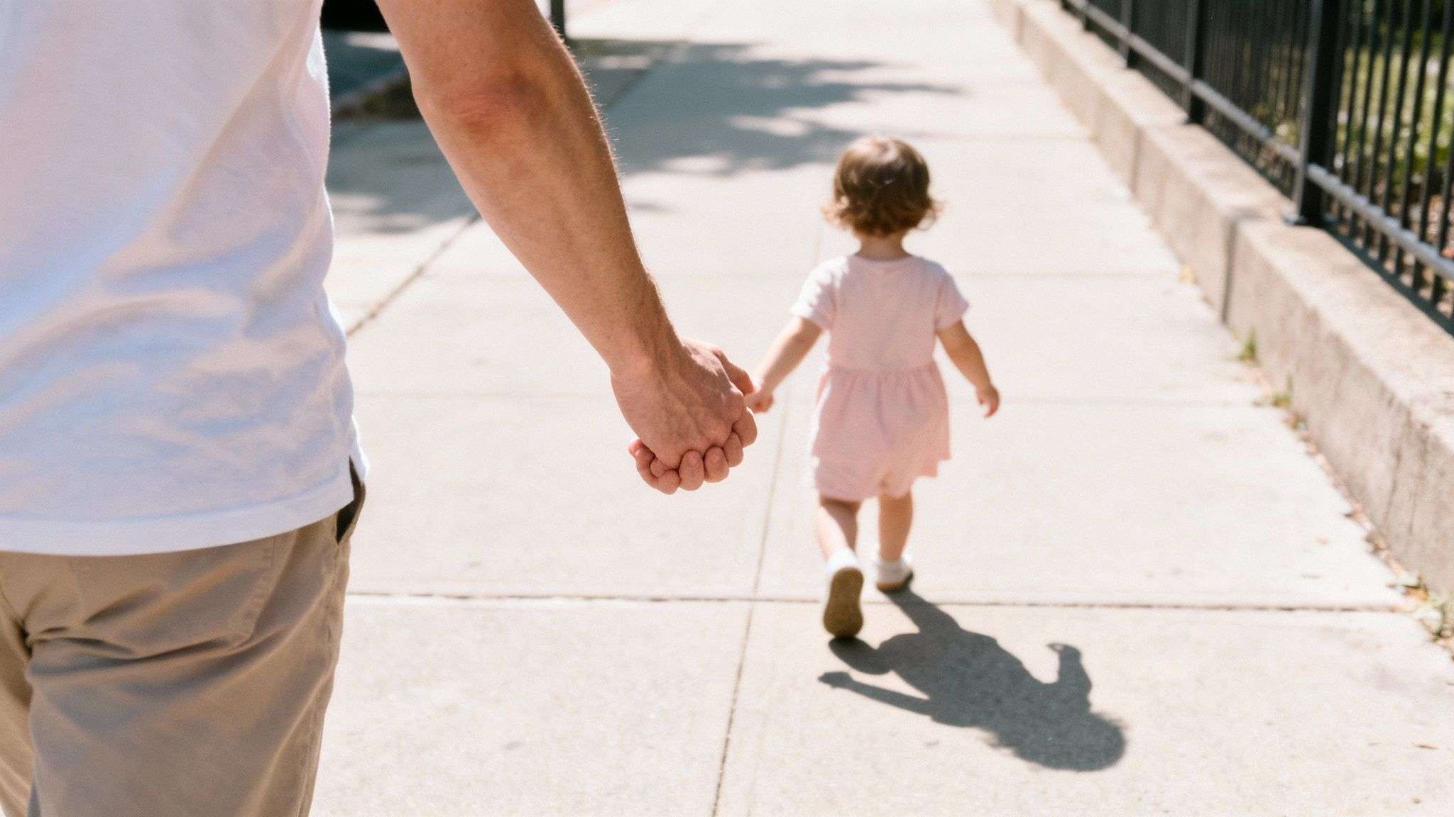 A parent holds a small child's hand while walking together on a sunny sidewalk.