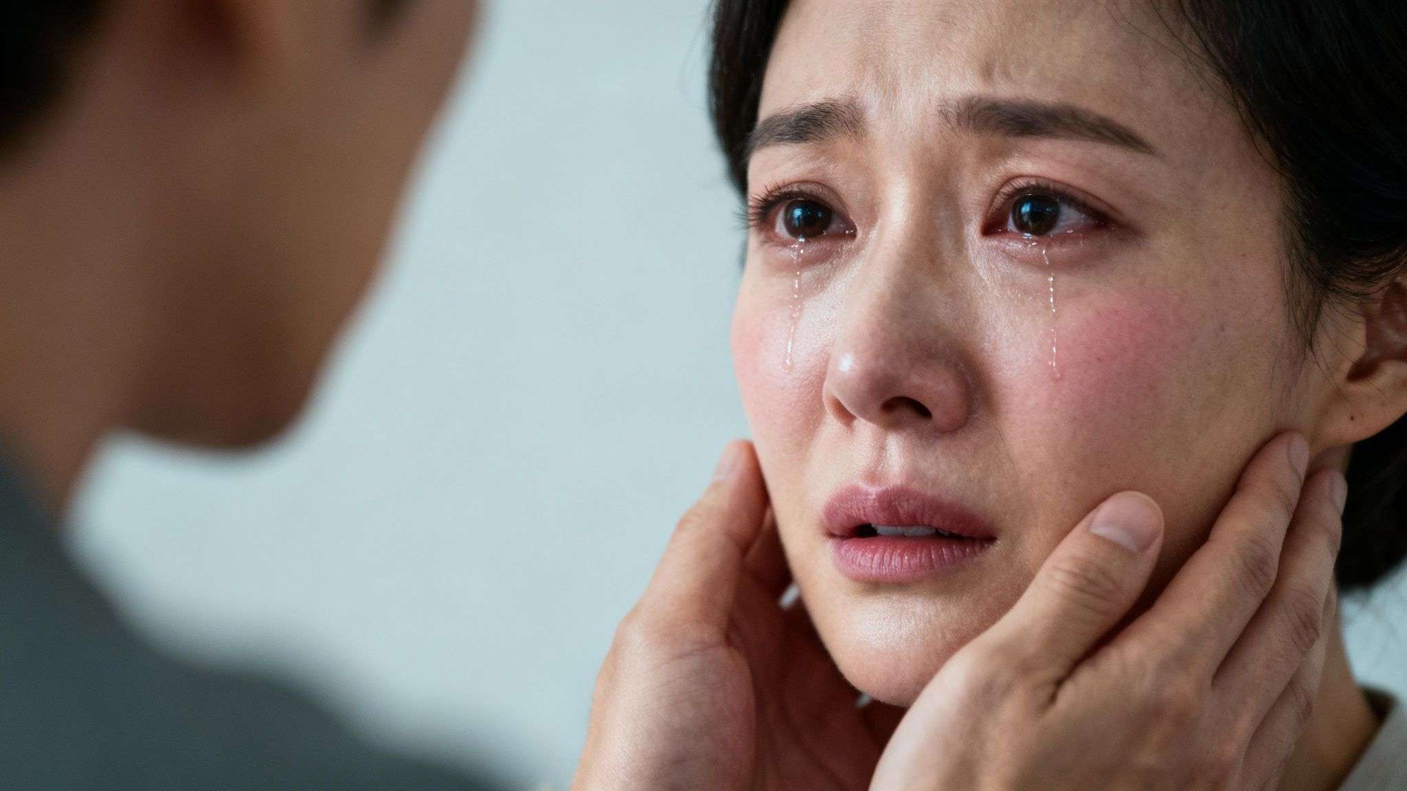 Close-up of a distraught woman crying, with tears streaming, as a man gently comforts her face.