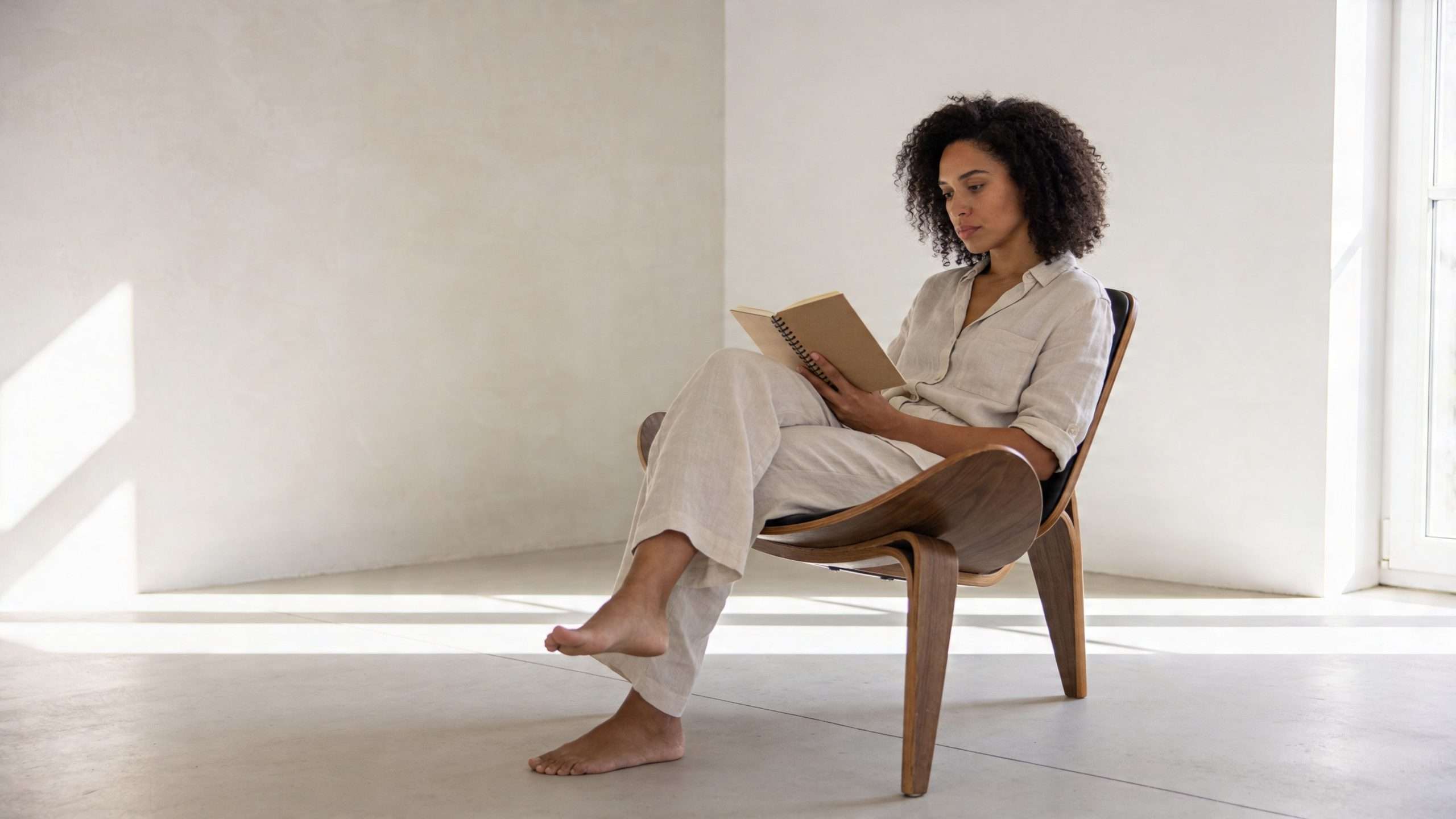 A woman with curly hair sitting in a wooden lounge chair reading a book in a minimalist room.