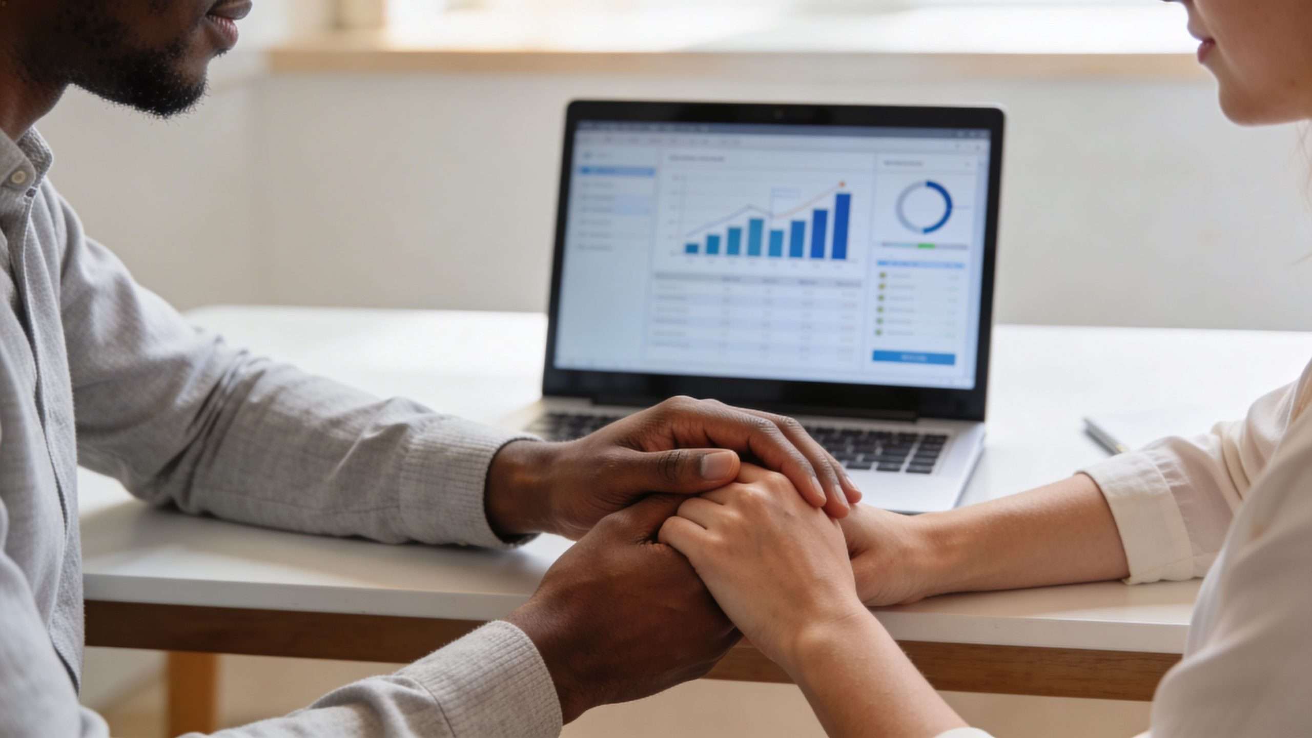 A couple holding hands while discussing financial charts on a laptop screen, emphasizing supportive communication about money.