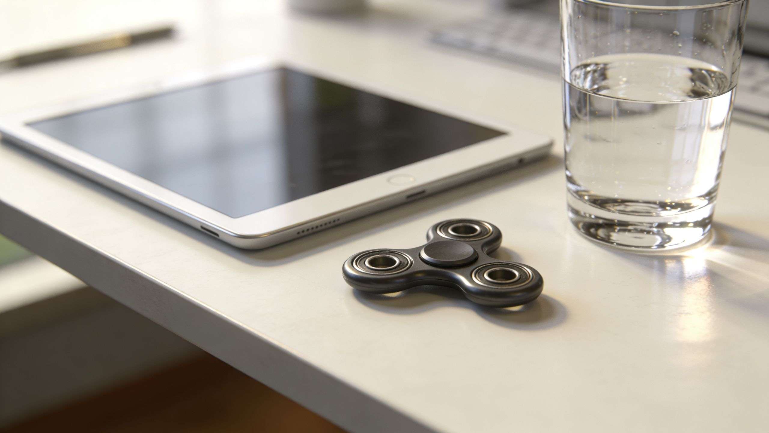 A minimalist office workspace featuring a tablet, a glass of water, and a metal fidget spinner.