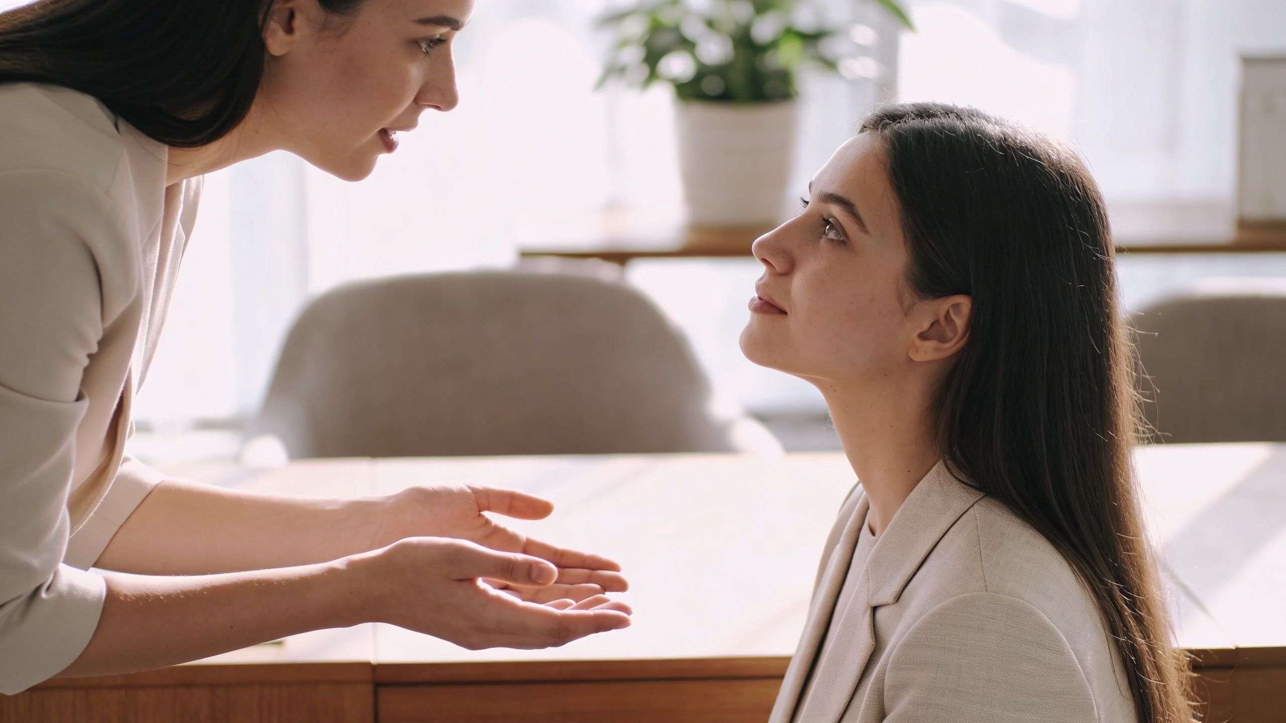 A professional woman attentively listens to a colleague who is speaking and gesturing in an office setting.