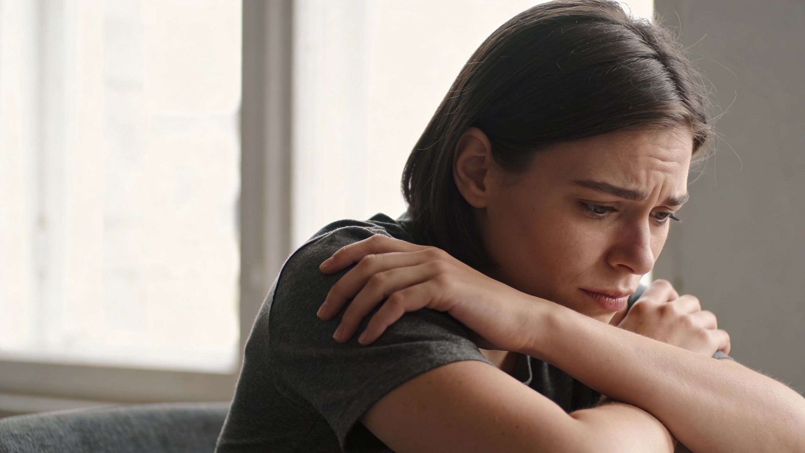 A close-up of a young woman appearing sad, thoughtful, and distressed while sitting and hugging herself.