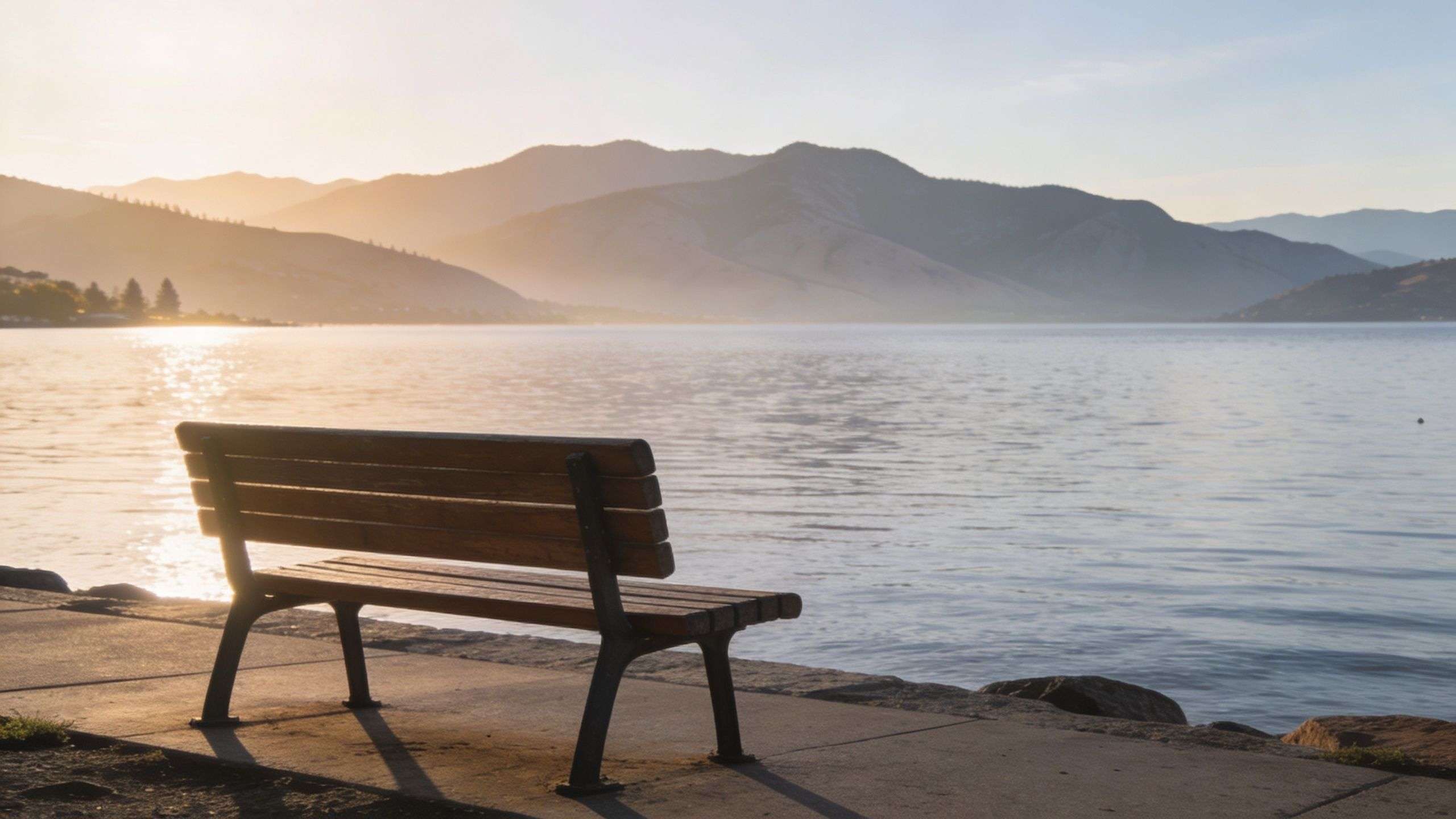 A lone wooden park bench overlooking a serene lake with mountains in the background during sunset.